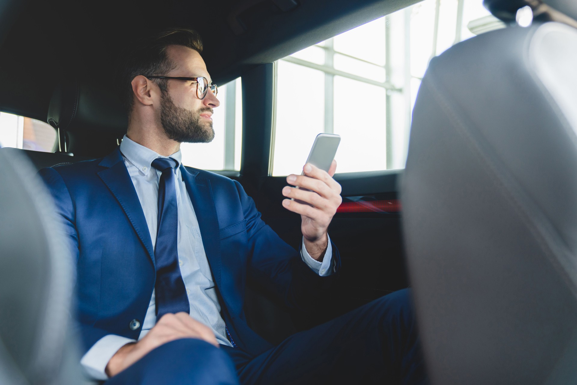 Confident successful rich young caucasian businessman in formal clothes using smart phone while sitting at backseat of taxi car with a driver looking at the window.